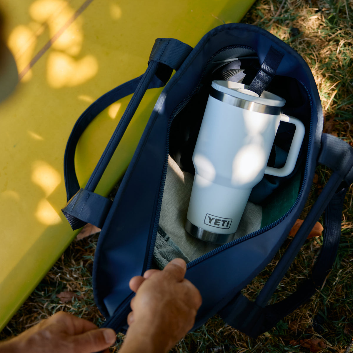 A navy blue tote bag is shown outdoors on grass with a hand opening the zipper. Inside the bag is a white Yeti travel tumbler with a stainless steel handle and lid.