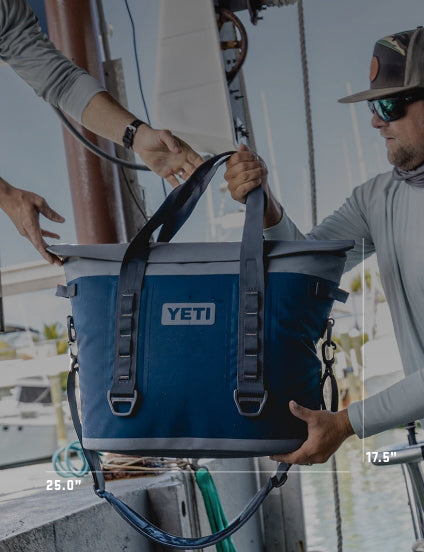 Two people holding a blue YETI cooler on a dock with water and boats in the background.