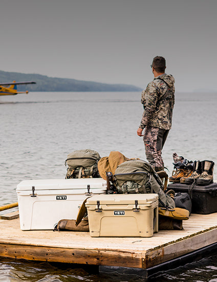 Person in camouflage clothing standing on a dock with YETI coolers and gear.