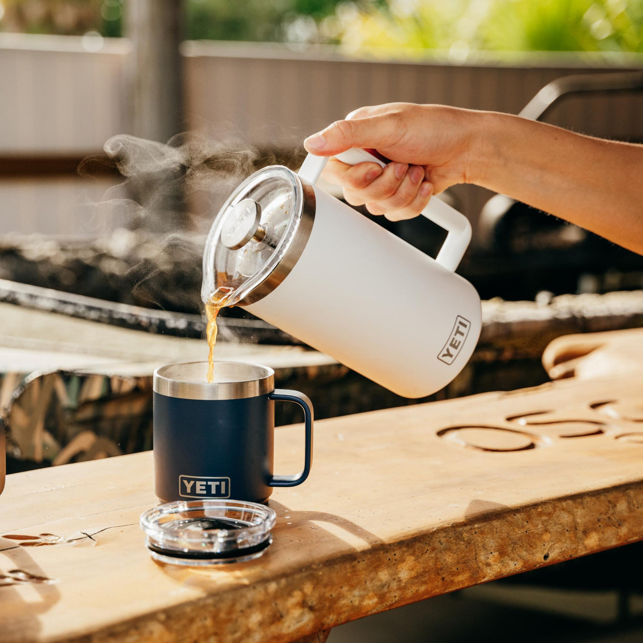 Person pouring coffee from a YETI french press into a YETI mug on a wooden table outdoors.