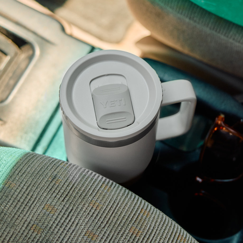 A white VETI travel mug with a handle and lid, shown sitting in a car cup holder next to sunglasses.