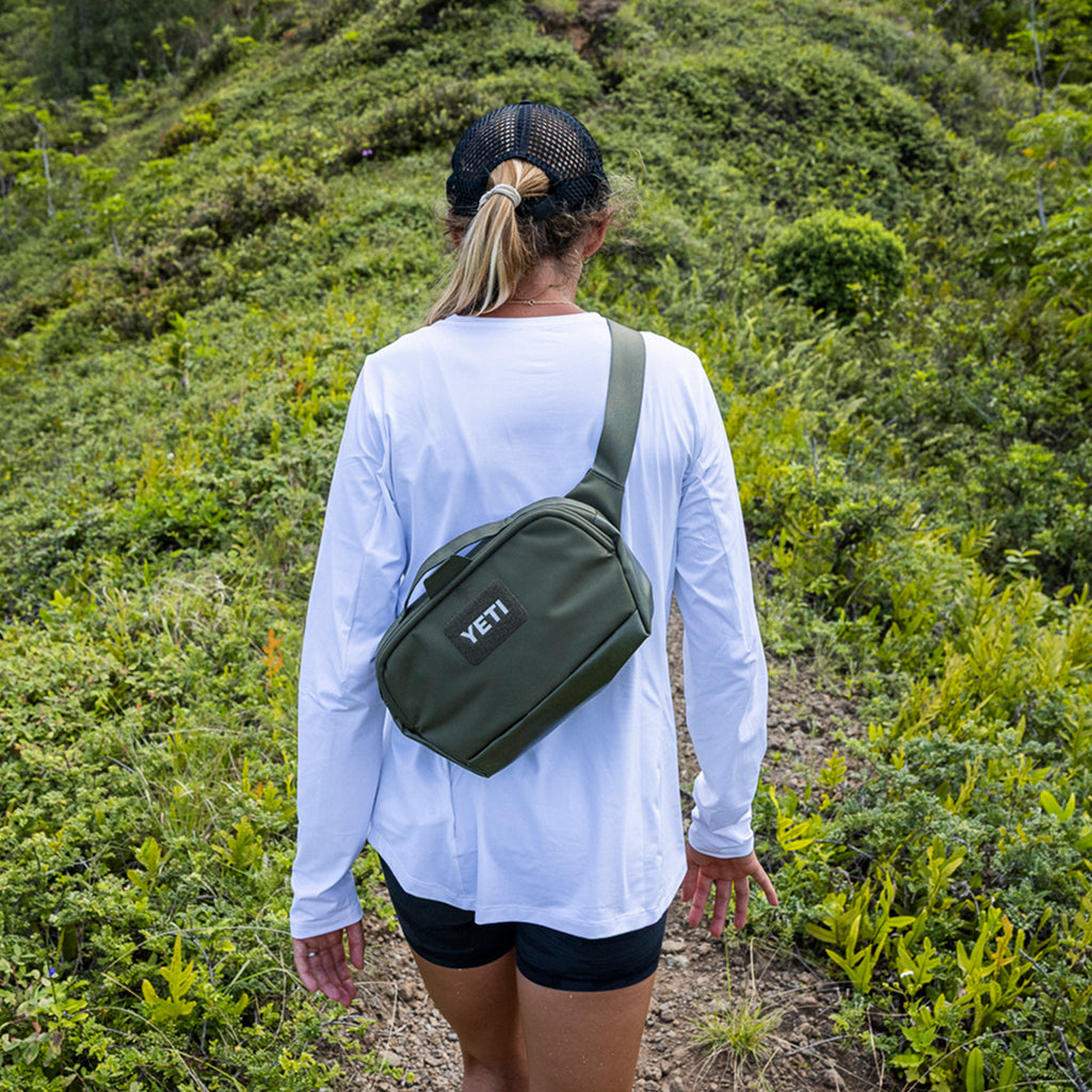 A woman viewed from behind wearing a white long-sleeve shirt and black shorts, carrying an olive green YETI crossbody bag on a trail with green vegetation in the background.