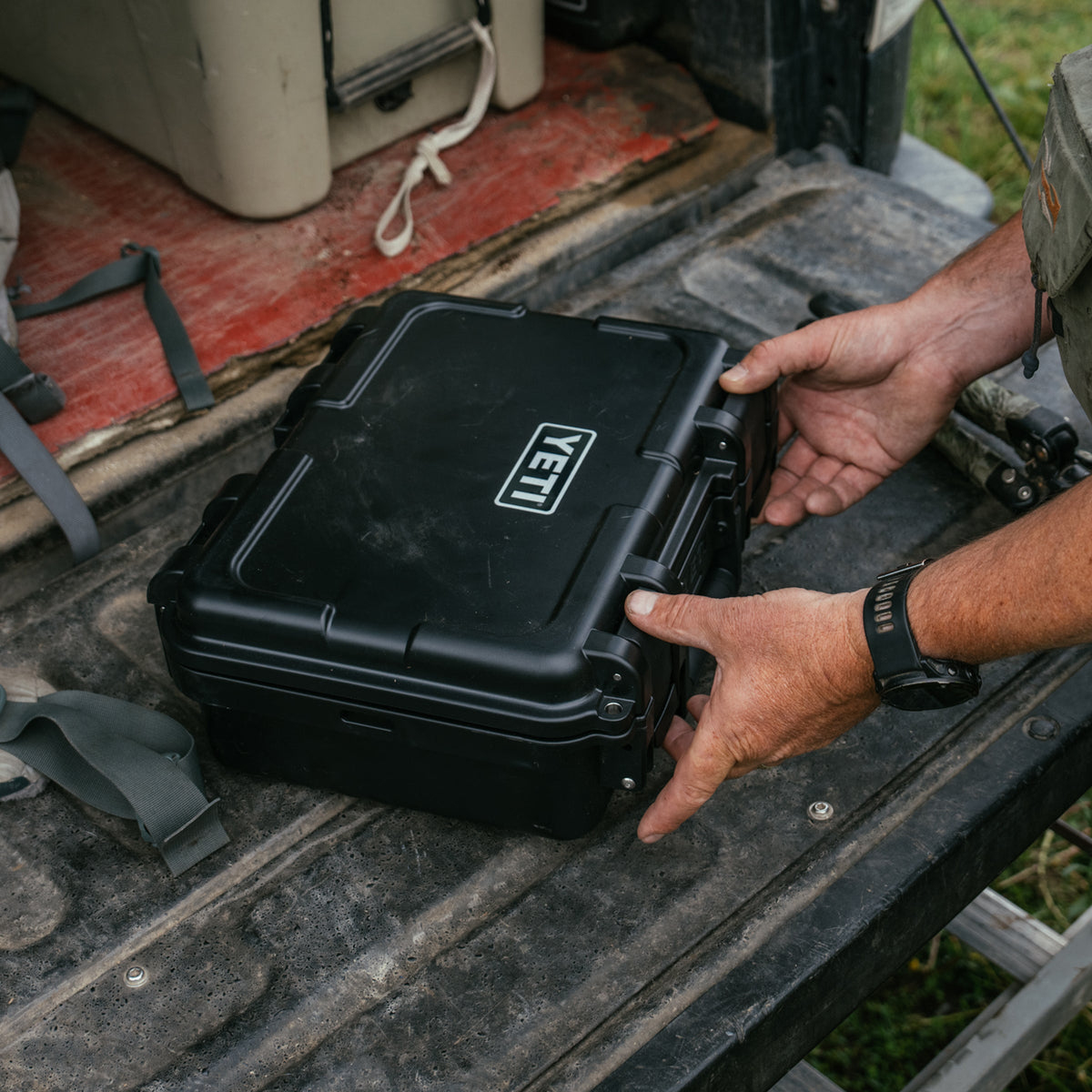 A black YETI hard case with the YETI logo on the lid, being held open by a person's hands on the tailgate of a truck.