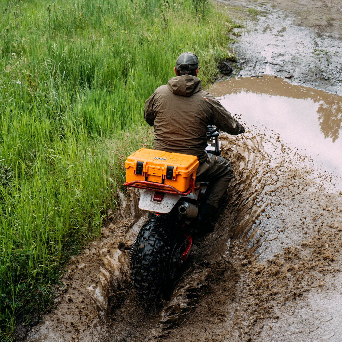 A person riding a motorcycle through a muddy puddle on a grassy path, viewed from behind. The motorcycle has a bright orange hard case mounted on the back.
