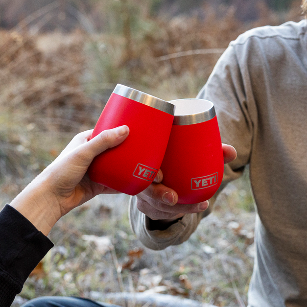 Two people sitting outdoors, holding red wine tumblers, with a bottle in the background.